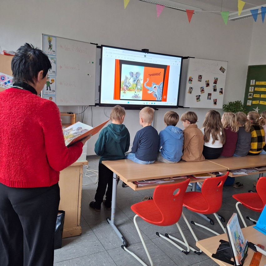 Eine Frau in einem roten Pullover liest ein Buch vor einer Gruppe von Kindern, die auf einer Bank im Klassenzimmer sitzen. Ein Monitor zeigt ein Bild eines Elefanten.