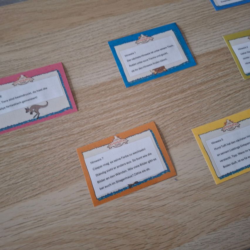 Ein Holztisch zeigt eine Reihe bunter Karten, jede mit unterschiedlichen Rändern. Eine Karte zeigt ein Känguru-Bild, eine andere einen Hasen und andere Karten haben Texte zu Tieren.