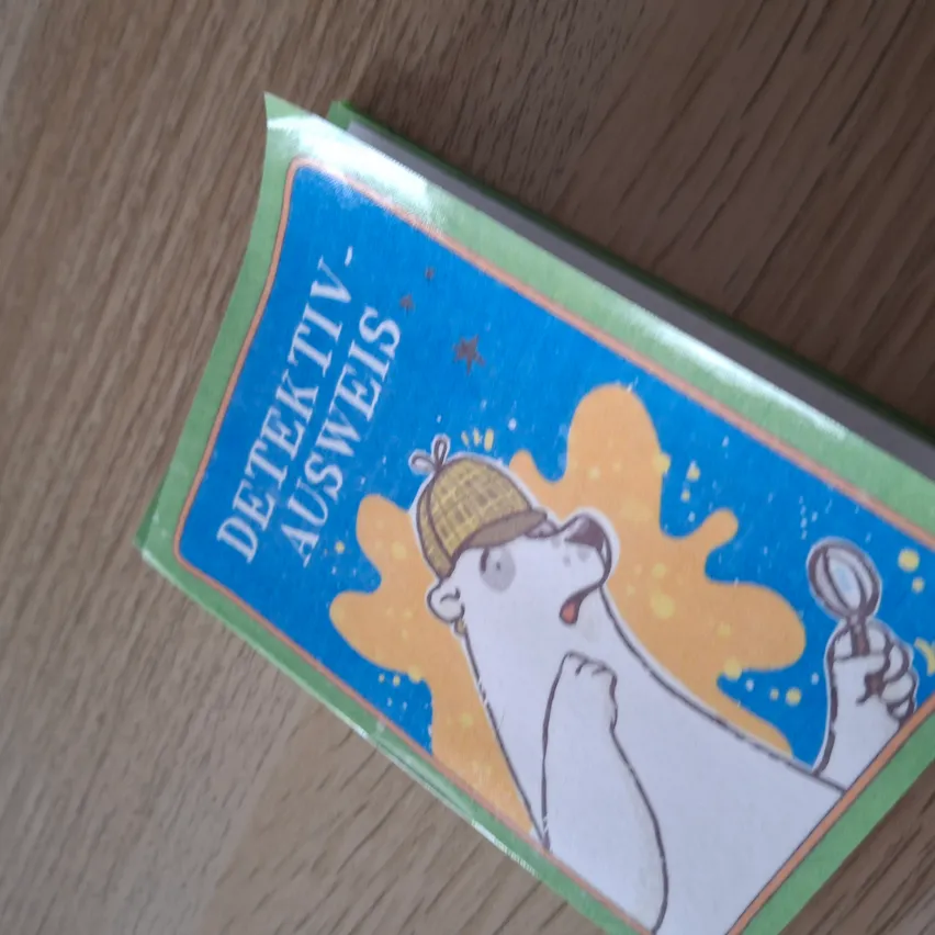 Ein Buch mit dem Titel 'Detektiv Ausweis' mit einem gezeichneten Eisbären in einem Detektivhut, der eine Lupe hält, und einige gelbe und blaue Spritzer auf einer Holzoberfläche.