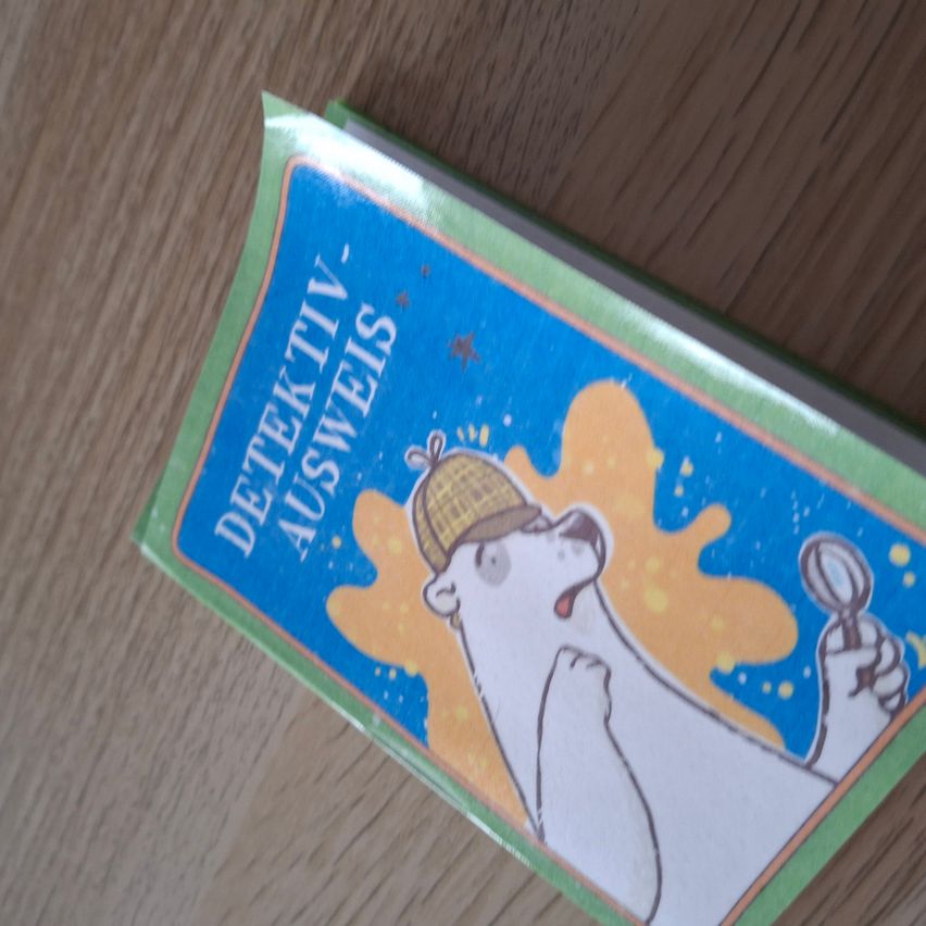 Ein Buch mit dem Titel 'Detektiv Ausweis' mit einem gezeichneten Eisbären in einem Detektivhut, der eine Lupe hält, und einige gelbe und blaue Spritzer auf einer Holzoberfläche.