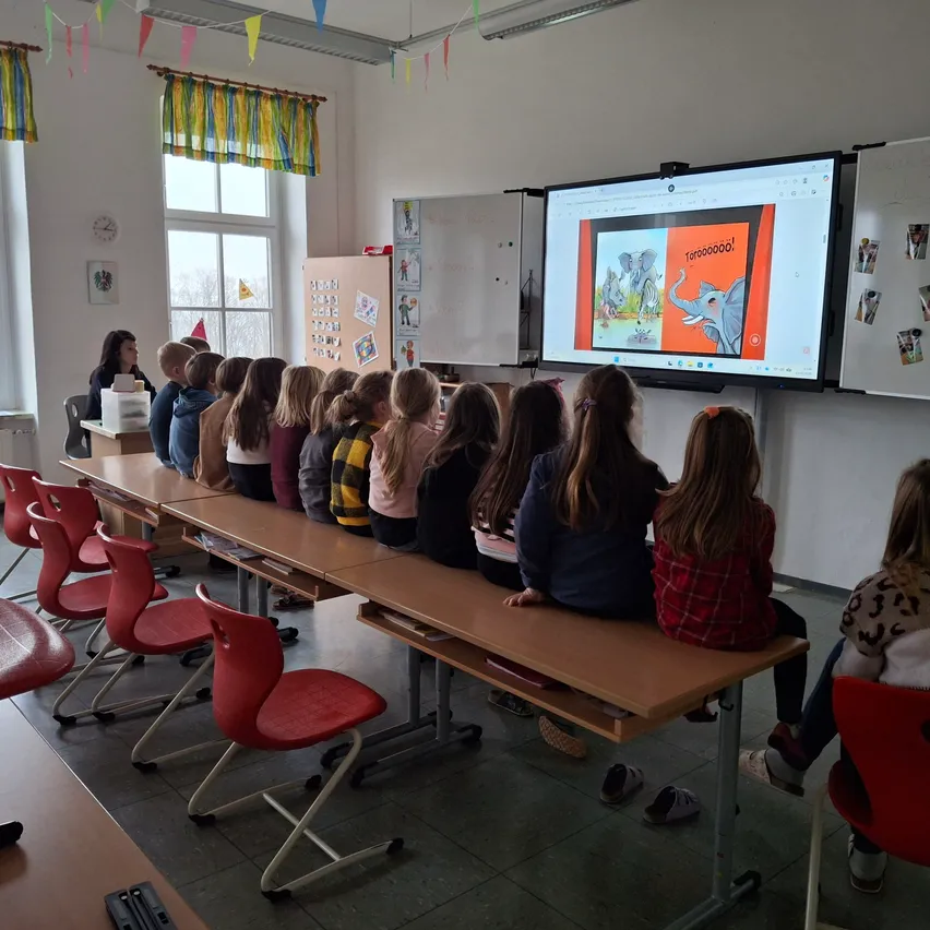Ein Klassenzimmer mit Schülern in Reihen, die eine Präsentation auf einem großen Bildschirm verfolgen. Bunte Dekorationen hängen an den Wänden, und eine Lehrerin ist im Hintergrund sichtbar.