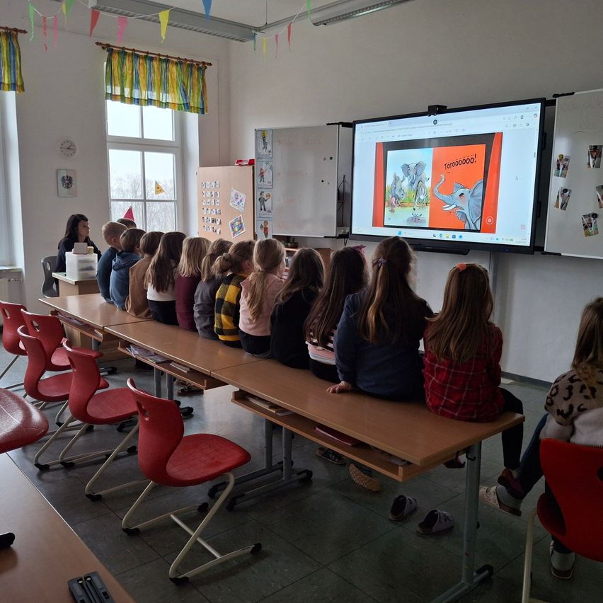Ein Klassenzimmer mit Schülern in Reihen, die eine Präsentation auf einem großen Bildschirm verfolgen. Bunte Dekorationen hängen an den Wänden, und eine Lehrerin ist im Hintergrund sichtbar.