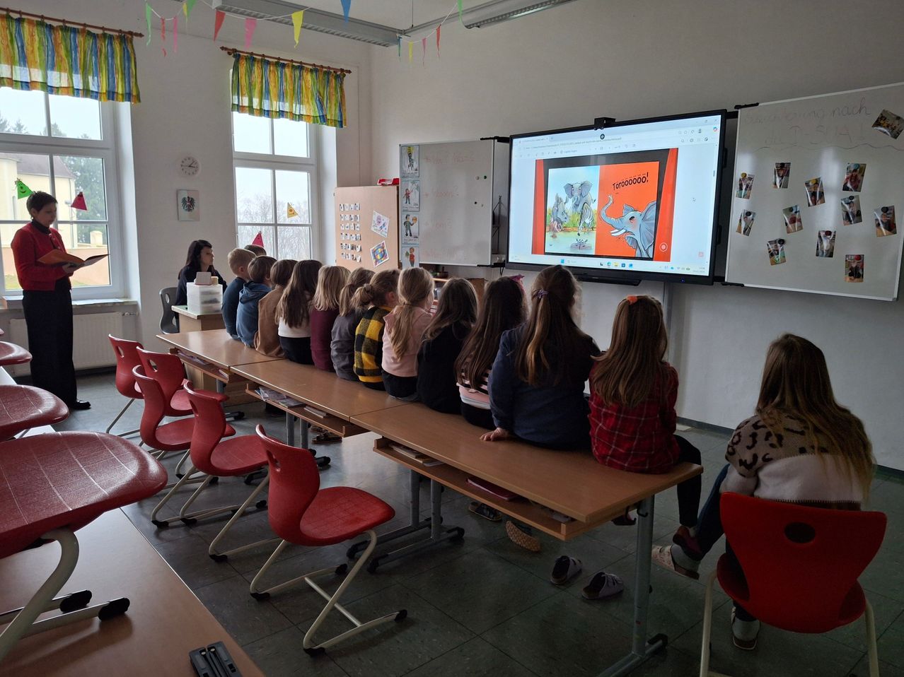 Ein Klassenzimmer mit Schülern in Reihen, die eine Präsentation auf einem großen Bildschirm verfolgen. Bunte Dekorationen hängen an den Wänden, und eine Lehrerin ist im Hintergrund sichtbar.
