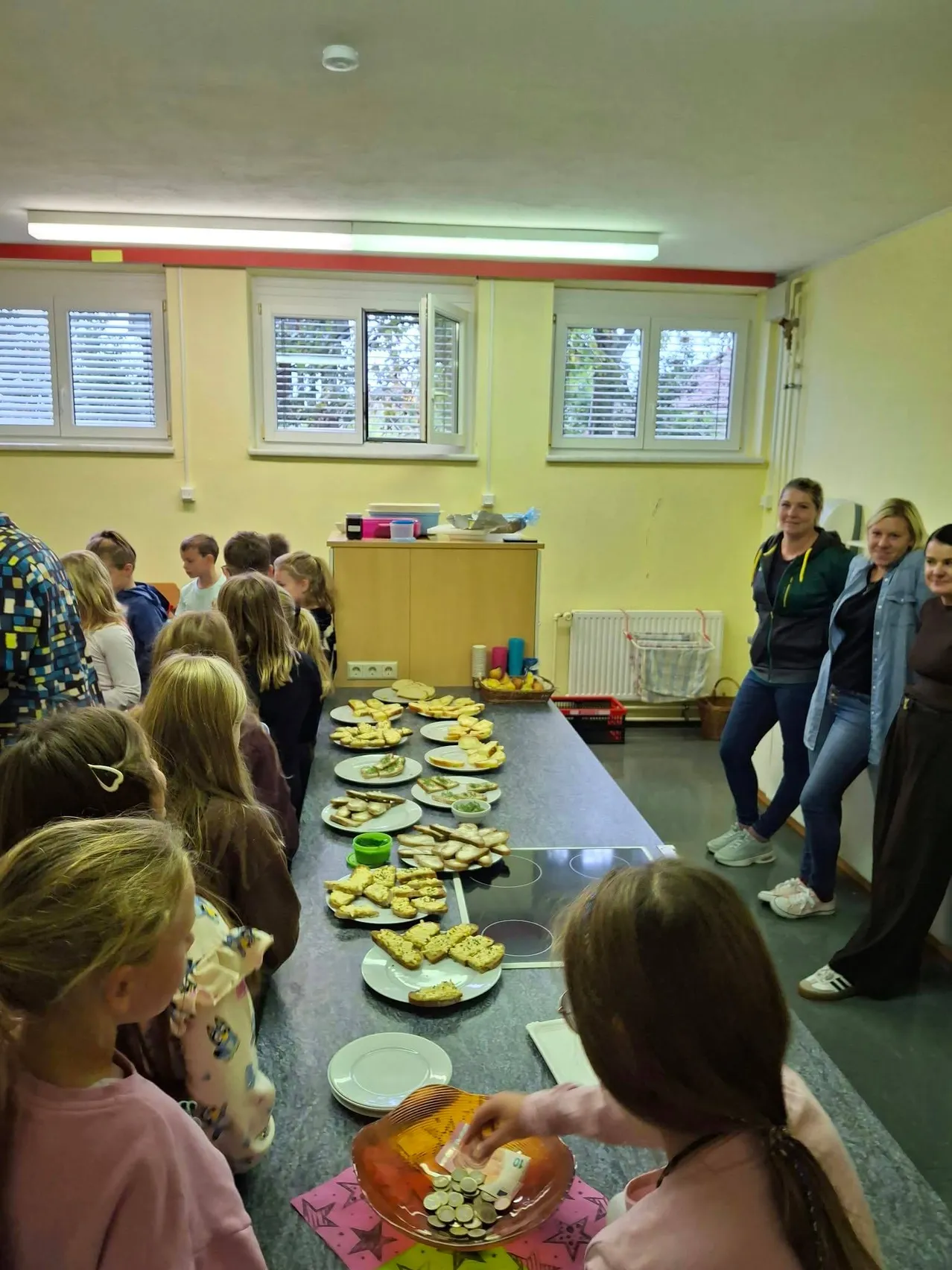 Eine Gruppe von Kindern und Erwachsenen versammelt sich um einen Tisch mit Essen in einem Raum.