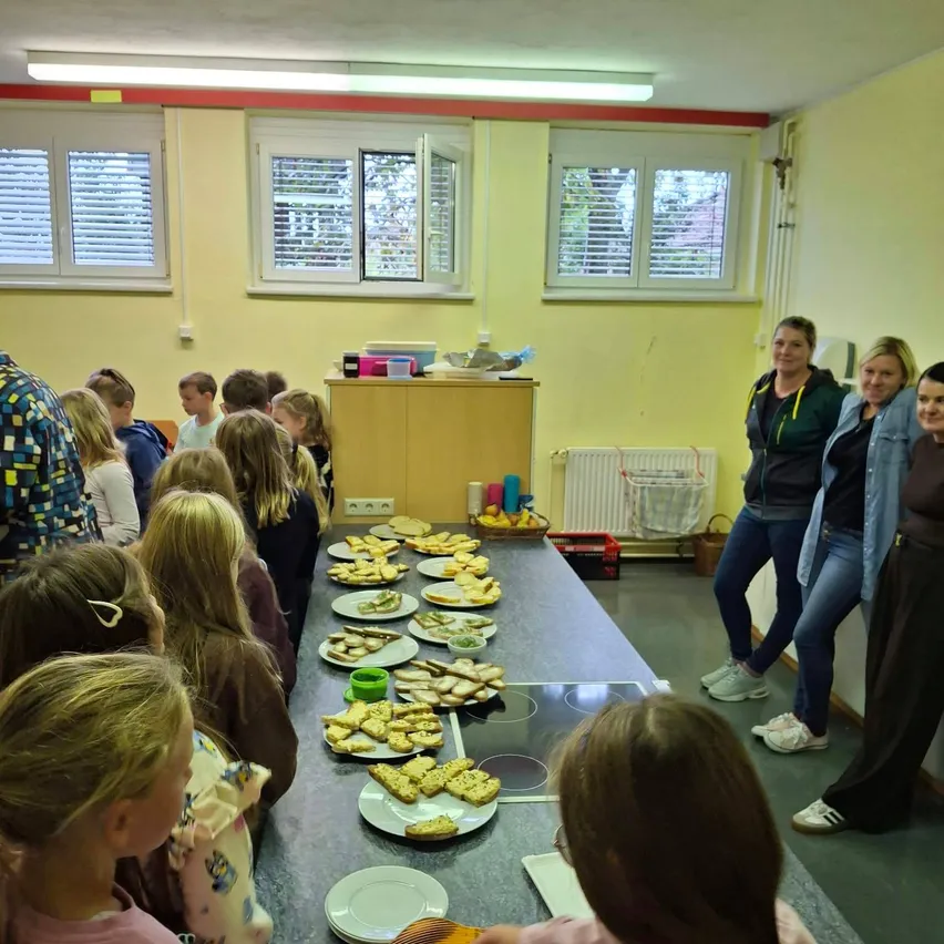 Eine Gruppe von Kindern und Erwachsenen versammelt sich um einen Tisch mit Essen in einem Raum.