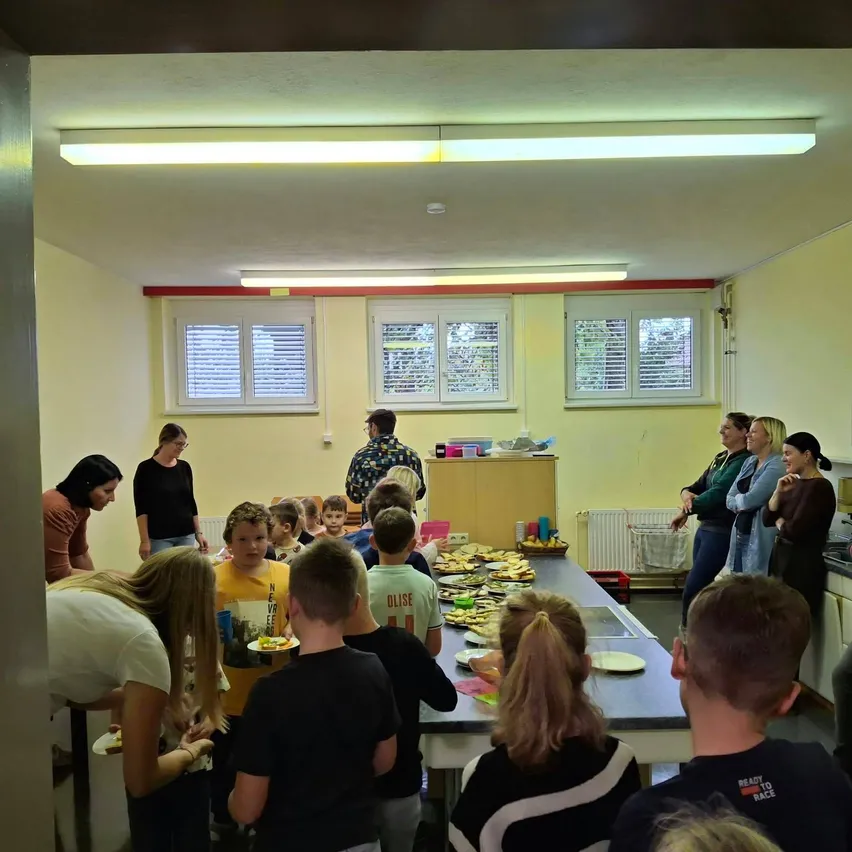 Eine Gruppe von Menschen, einschließlich Erwachsener und Kindern, versammelt sich in einem Raum. Sie stehen um einen Tisch herum, der mit Essen gefüllt ist. Einige halten Teller, während andere stehen und zusehen.