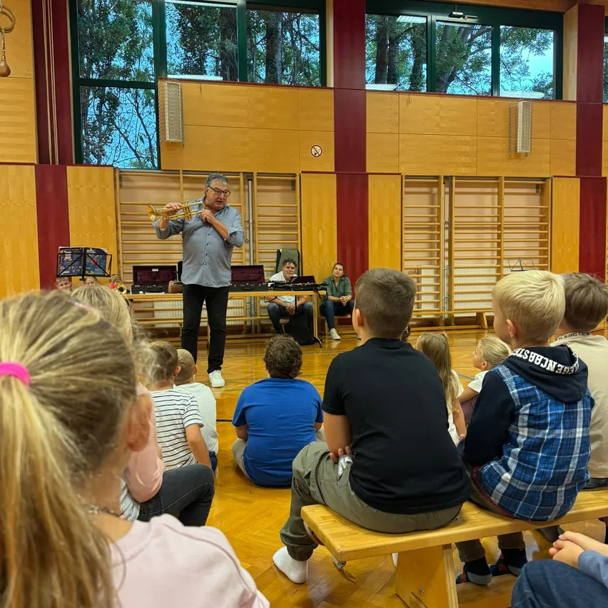 Ein Mann spielt Trompete in einem Raum, in dem Kinder auf Bänken sitzen.