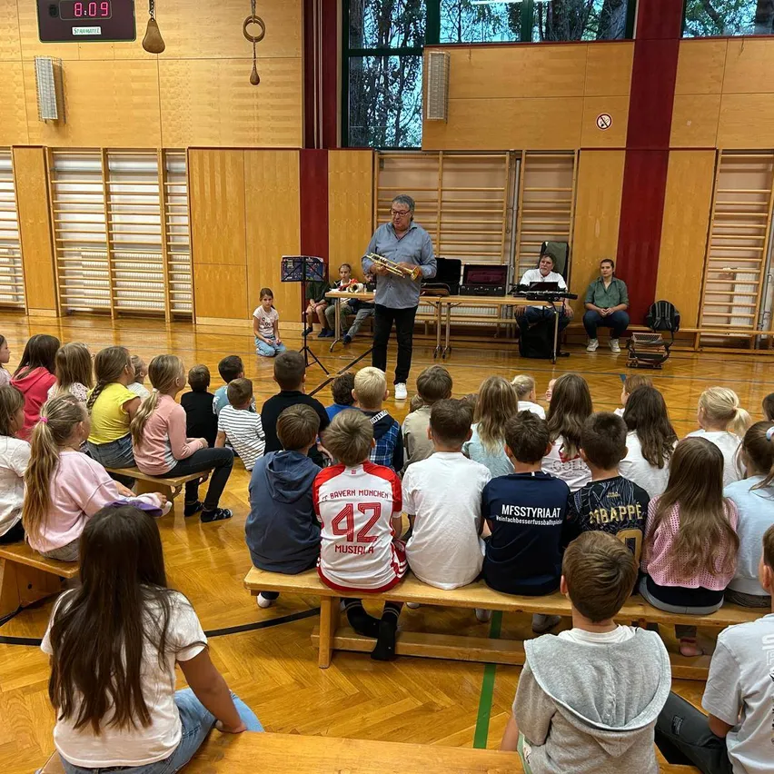 Ein Mann steht vor einer Gruppe von Kindern in einer Turnhalle, hält eine Trompete und liest von einem Notenständer.