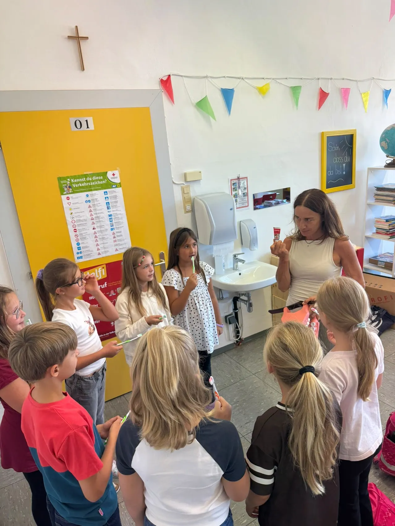 Eine Gruppe von Kindern versammelt sich in einem Klassenzimmer, sie halten Zahnbürsten und Zahnpasta. Eine Frau steht vor ihnen und hält eine Zahnbürste und Zahnpasta.