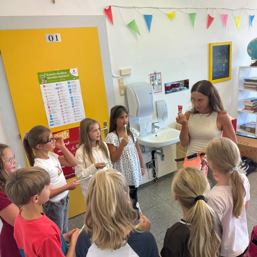 Eine Gruppe von Kindern versammelt sich in einem Klassenzimmer, sie halten Zahnbürsten und Zahnpasta. Eine Frau steht vor ihnen und hält eine Zahnbürste und Zahnpasta.