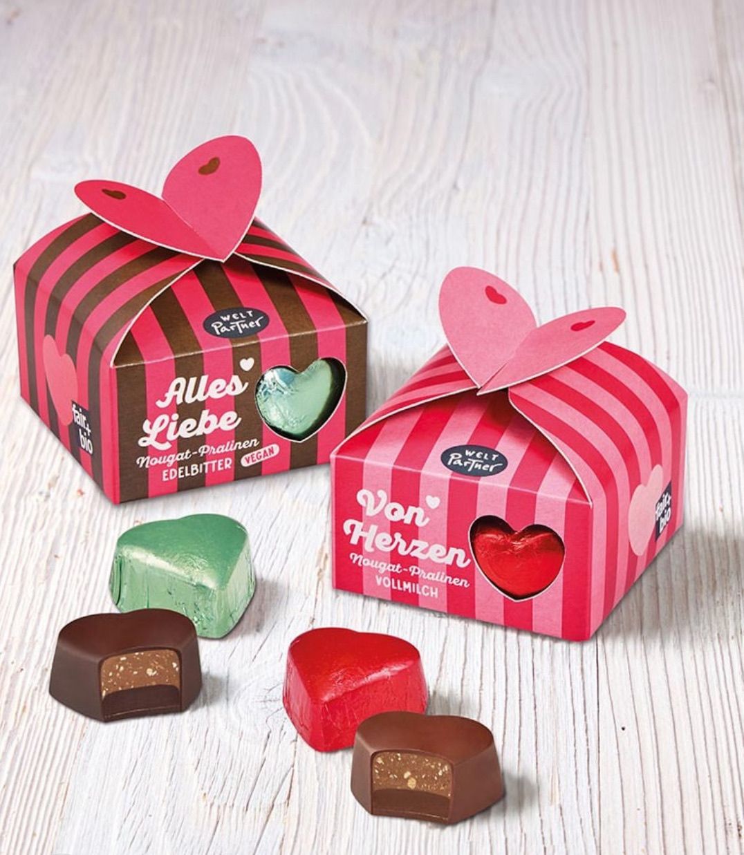Zwei rosa Geschenkboxen mit herzförmigen Deckeln, beschriftet mit 'Alles Liebe' und 'Von Herzen', stehen auf einer Holzoberfläche. Darunter befinden sich verschiedene Schokoladen in Herzform, einschließlich grün, rot und braun.