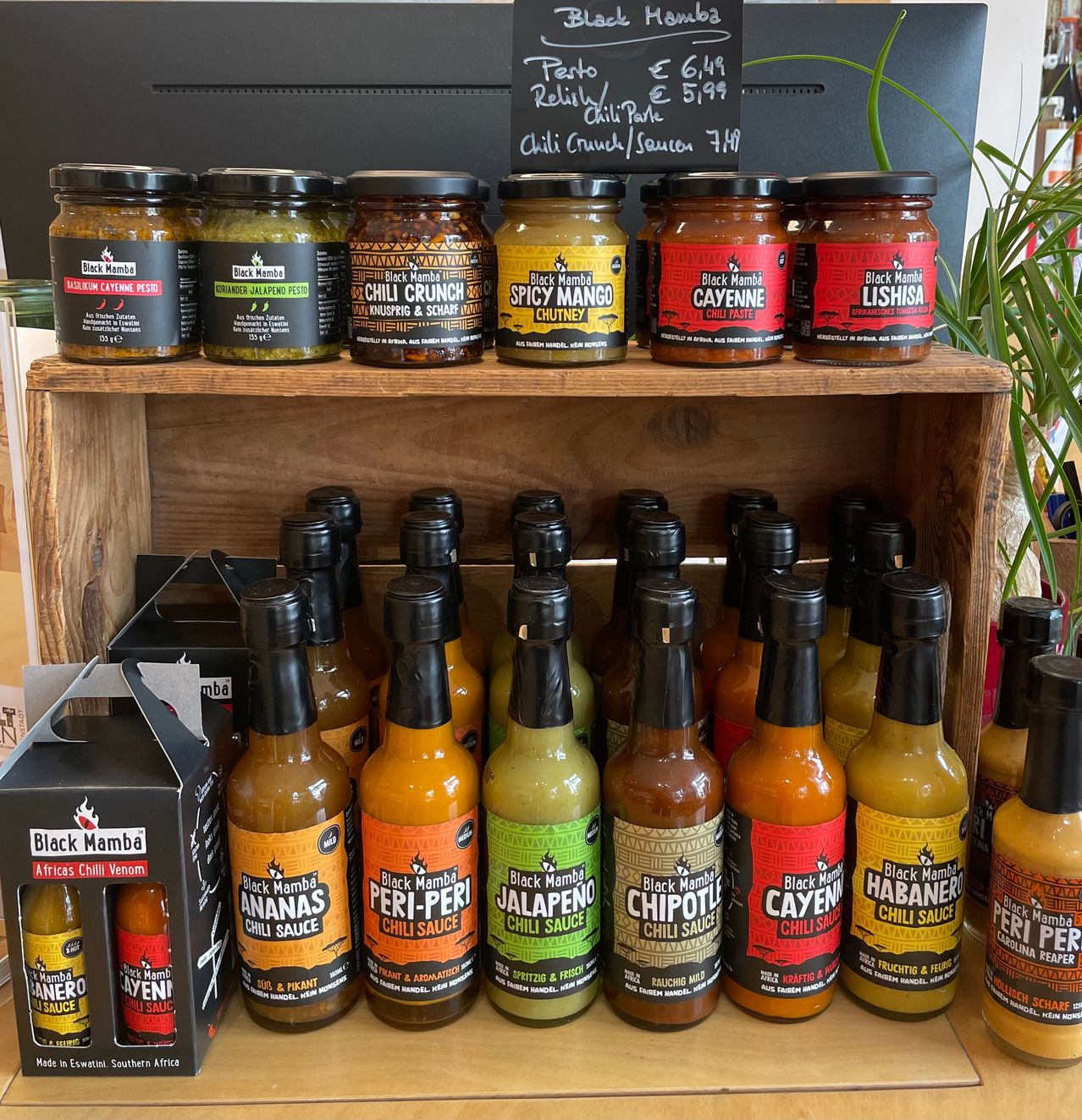 Ein hölzerne Ablage zeigt verschiedene Flaschen der Black Mamba Chilisaucen, einschließlich Geschmacksrichtungen wie Cayenne, Chili Crunch und Spicy Mango.