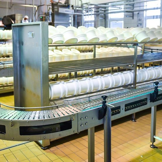 Eine Fabrik mit mehreren Regalen, die mit weißen Behältern auf einem Förderband gestapelt sind. Eine Person steht hinter der Maschine.