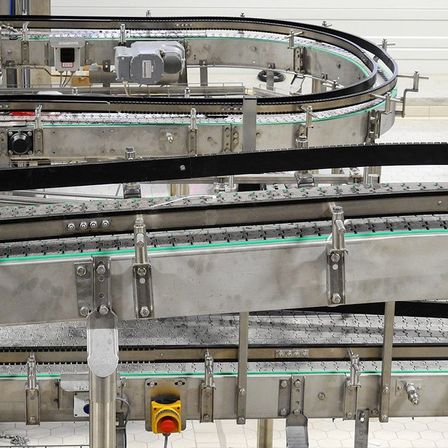 Ein Förderbandsystem mit mehreren Metallschienen, Rollen und Halterungen ist abgebildet. Schalter und ein gelber Knopf sind am Förderband zu sehen.
