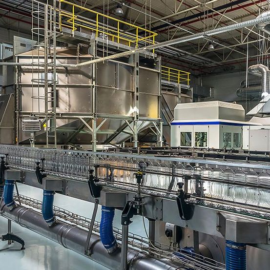 Eine Produktionsanlage mit einem Förderbandsystem und großen Maschinen im Betrieb. Das Förderband hat sich bewegende Glasflaschen. Oben gibt es Metallgerüste und Rohre.