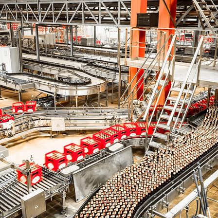 Eine große Fabrik mit mehreren Förderbändern und roten Kisten darauf. Eine Treppe und Metallgeländer befinden sich auf der rechten Seite.