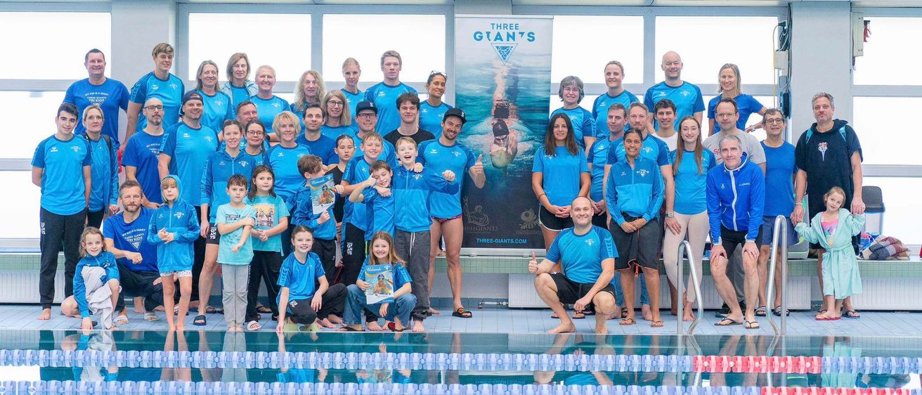 Eine Gruppe von Menschen in blauen Shirts und Badekleidung steht am Pool mit einem Poster eines Schwimmers. Einige lächeln, und ein Mann kniet mit Daumen hoch.