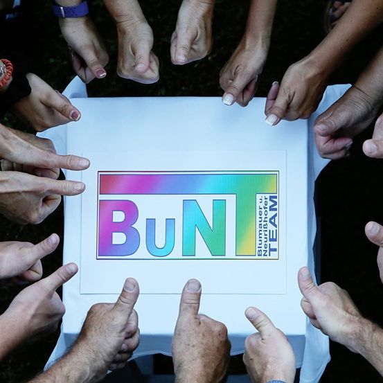 Hände berühren eine weiße Box mit dem Text BuN darauf. Die Box befindet sich in der Mitte der Hände. Die Hände zeigen auf die Box.
