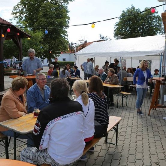 Menschen versammeln sich draußen für eine Veranstaltung. Einige sitzen an Tischen unter Zelten, andere stehen herum. Bäume und Häuser sind im Hintergrund.
