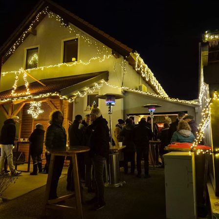 Eine Gruppe von Menschen steht bei Nacht vor einem Haus mit Weihnachtsbeleuchtung. Die Menschen stehen um Tische herum.