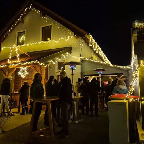 Eine Gruppe von Menschen steht bei Nacht vor einem Haus mit Weihnachtsbeleuchtung. Die Menschen stehen um Tische herum.