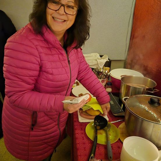 Eine Frau in einer rosa Jacke steht in einer Küche, hält Essen und lächelt. Ein Tisch hinter ihr ist mit Schüsseln, Löffeln und einem Topf gedeckt.