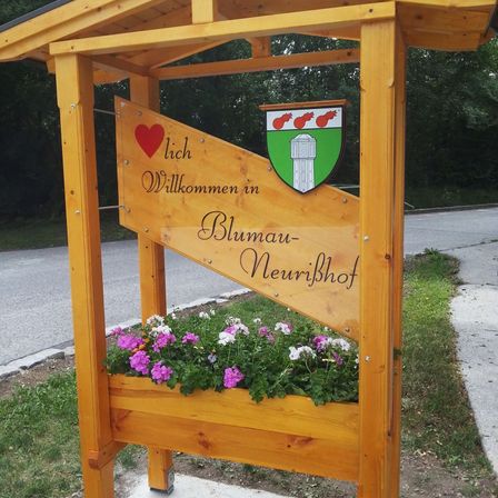 Ein hölzernes Schild mit Blumen heißt Sie in Blumau-Neurihof willkommen. Das Schild zeigt ein Herz und das Wappen des Dorfes. Bäume und eine Straße sind im Hintergrund zu sehen.