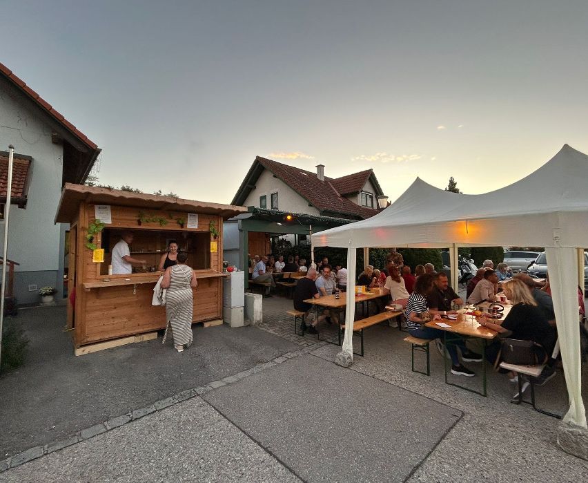 A bustling outdoor dining area with a wooden food stand and a white tent, filled with people enjoying their meals under the evening sky.