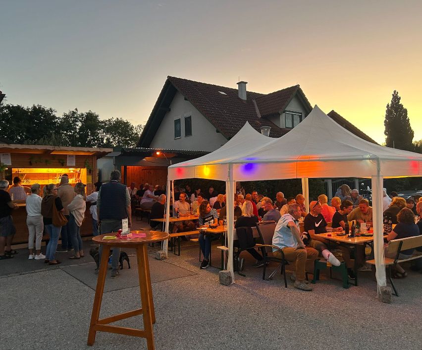 A group of people are gathered around tables under a tent, enjoying drinks and food in an outdoor setting. The sky is orange, and there is a house in the background.