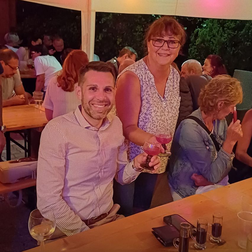 A group of people are gathered at an outdoor event, smiling and holding drinks. A man and a woman stand in the foreground, smiling and holding glasses of pink liquid.