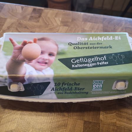 Eine Packung Aichfeld-Eier aus der Obersteimark auf einer Holzoberfläche. Die Packung enthält 10 Eier, freilandgehegt und ohne Gentechnik.