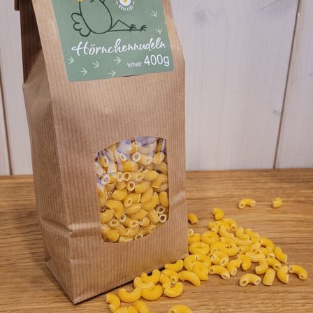 Ein brauner Papierbeutel mit einem grünen Aufkleber, der eine Henne zeigt, beschriftet mit 'Hörchennudeln' und 'Inhalt: 400g', gefüllt mit Nudeln, mit einigen Nudeln auf einem Holztisch verstreut.
