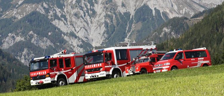 Vier Feuerwehrfahrzeuge und ein Van sind auf einem Grashügel geparkt, mit einem Gebirgszug im Hintergrund.