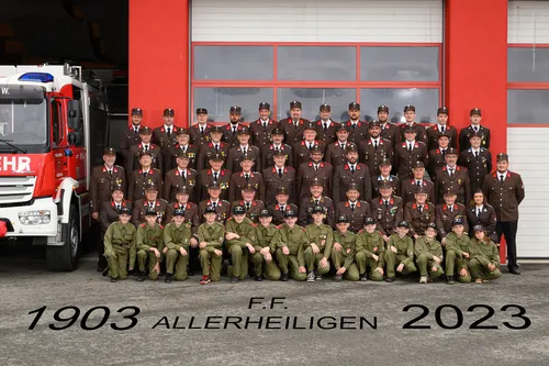 Gruppenfoto von Feuerwehrleuten in grünen Uniformen, die vor einem roten Gebäude posieren. Der Text '03 Allerheiligen 20' steht unten.