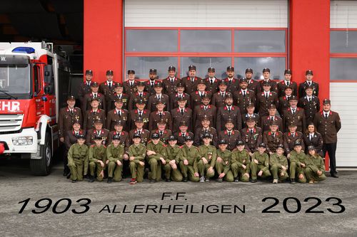 Gruppenfoto von Feuerwehrleuten in grünen Uniformen, die vor einem roten Gebäude posieren. Der Text '03 Allerheiligen 20' steht unten.
