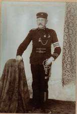 Ein altes Foto eines Mannes in Militäruniform, neben einem behangenen Tisch stehend. Er hält ein Schwert und hat Medaillen auf der Brust.