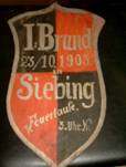 Ein Vintage-Abzeichen mit dem Text 'I. Brand 25/10/1908 Siebing' und dem Logo einer Feuerwehr. Das Abzeichen ist rot, schwarz und weiß, mit dem Datum in der Mitte.