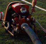 Ein roter Bestandteil einer Feuerwehrspritze liegt auf einem Rasenfeld, mit einem Schlauch angeschlossen. Eine Hand eines Menschen ist sichtbar, möglicherweise justiert oder überprüft die Ausrüstung.