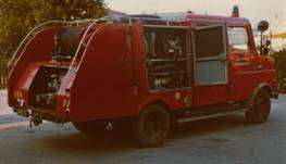 Ein roter Feuerwehrwagen im Vintage-Stil mit offenen Hecktüren steht auf einem Betonweg, umgeben von Bäumen.