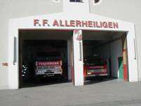Eine Feuerwehrstation namens F.F. Allerheiligen mit zwei geparkten Feuerwehrwagen, einer mit der Aufschrift 'Feuerwehr'. Die Station hat eine weiße Außenseite und rote Verzierungen.