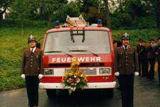 Zwei Feuerwehrleute stehen vor einem Feuerwehrauto mit einem Kranz vorne. Dahinter steht eine Gruppe von Menschen in Uniform auf einer Rasenfläche.