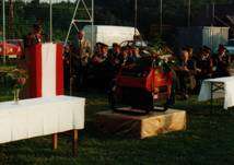 Eine Versammlung auf einem Feld mit einem Podium, Tischen und Menschen. Ein rotes Maschinenfahrzeug steht auf der Bühne. Bäume und andere Fahrzeuge sind im Hintergrund.