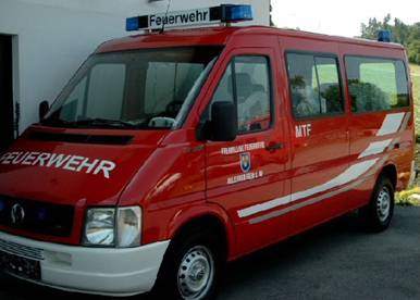 Ein rotes Notfallfahrzeug mit dem Wort Feuerwehr auf der Seite und einer blauen Lichtleiste oben. Das Fahrzeug ist vor einem Gebäude geparkt.
