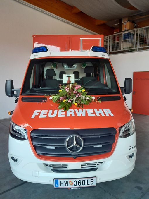 Ein orangefarbener Mercedes-Benz Feuerwehrwagen mit dem Wort 'FEUERWEHR' auf der Front steht in einer Garage. Im Inneren befinden sich zwei schwarze Stühle, und ein Blumenstrauß schmückt den vorderen Stoßfänger.