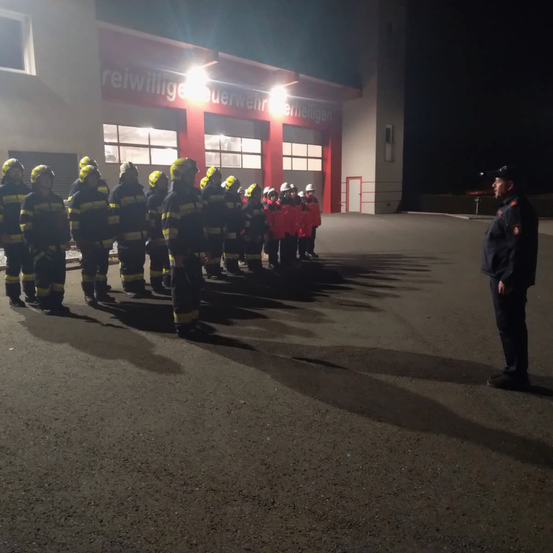 In der Nacht steht eine Gruppe von Feuerwehrleuten in Formation vor einer Feuerwache. Ein Offizier spricht sie an.