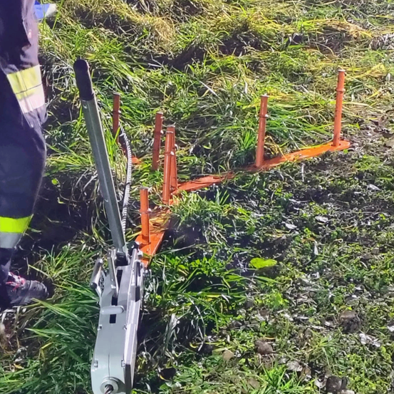 Ein Feuerwehrmann in voller Ausrüstung steht neben einem langen orangefarbenen Rahmen mit mehreren Stangen, möglicherweise einem Feuerwehrwerkzeug, auf einem Grasfeld.