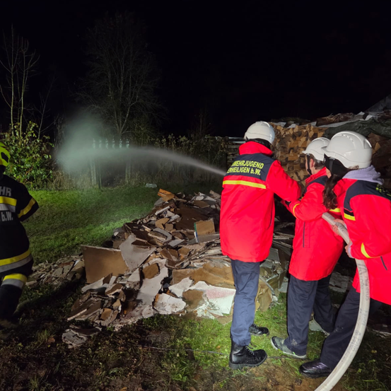 Feuerwehrleute verwenden einen Schlauch, um einen Haufen Schutt auf einem Rasenfeld bei Nacht zu löschen.