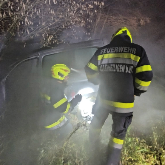 Zwei Feuerwehrleute in Schutzkleidung retten jemanden aus einem Fahrzeug. Die Szene ist rauchig und neblig.