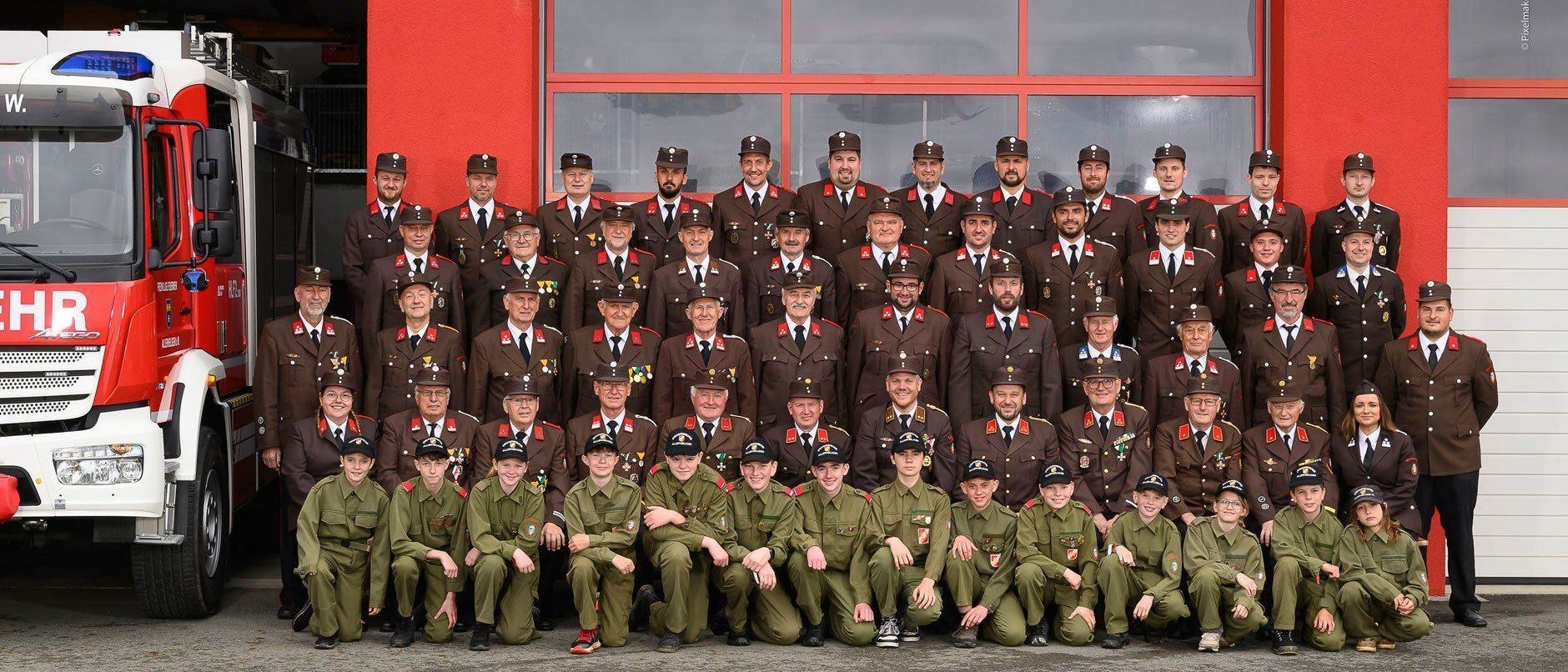 Ein Gruppenfoto von uniformierten Personen steht vor einem roten Gebäude. Sie tragen Mützen und haben Abzeichen an ihren Uniformen. Es gibt zwei Reihen, eine kniend und eine stehend. Der Text lautet '03 Allerheiligen 20'.
