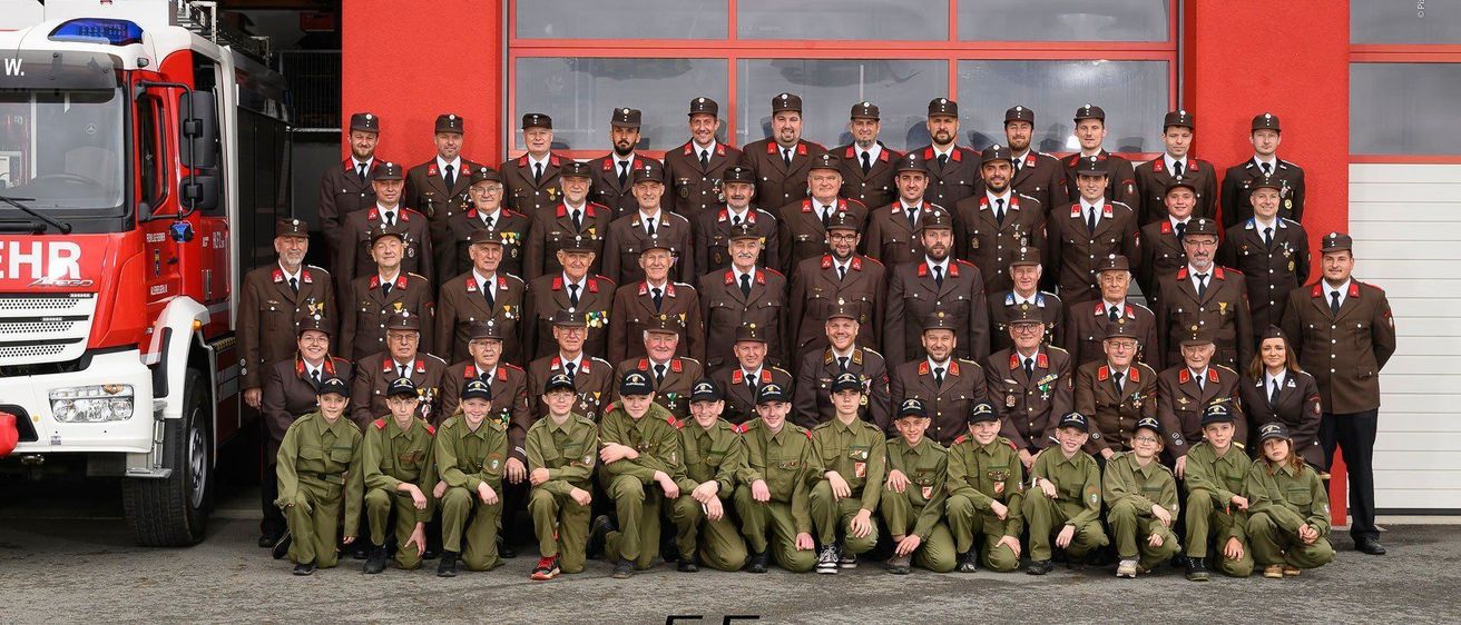 Ein Gruppenfoto von uniformierten Personen steht vor einem roten Gebäude. Sie tragen Mützen und haben Abzeichen an ihren Uniformen. Es gibt zwei Reihen, eine kniend und eine stehend. Der Text lautet '03 Allerheiligen 20'.